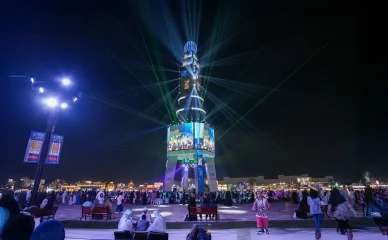 برج احتفالي مضاء يعرض شاشات وأشعة ليزر خضراء وسط حشد من الزوار ليلاً.