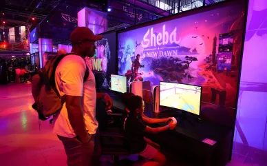 رجل يقف حاملاً حقيبة ظهر ويشاهد طفلاً جالساً يلعب لعبة الفيديو "Sheba: A New Dawn" على شاشة عرض داخل قاعة معارض مضاءة بألوان النيون الوردية والبنفسجية.