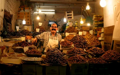 بائع مبتسم يقف خلف منصة عرض كبيرة مليئة بأكوام متنوعة من التمور داخل متجر مضاء بمصابيح دافئة في سوق شعبي.