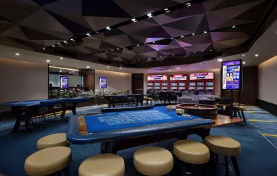 صالة كازينو حديثة تحتوي على طاولات لعب زرقاء وماكينات قمار تحت سقف بتصميم هندسي.