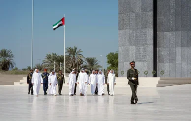 "مسؤولون إماراتيون في كندورة بيضاء وعسكريون في زي عسكري يسيرون معاً كوفد رسمي على ساحة بيضاء واسعة. في الخلفية علم دولة الإمارات يرفرف على