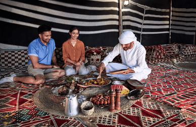 رجل إماراتي يرتدي الزي التقليدي يقدم وجبة لضيفين سائحين في خيمة بدوية تراثية. الضيفان ينظران إلى الأطباق التقليدية المتنوعة على السجادة الملونة.