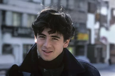 Jeune Patrick Bruel dans la rue, vêtu d’un manteau noir.