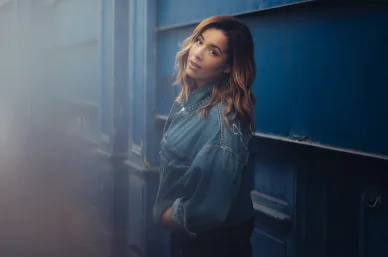 Chimène Badi en veste en jean posant contre un mur bleu.