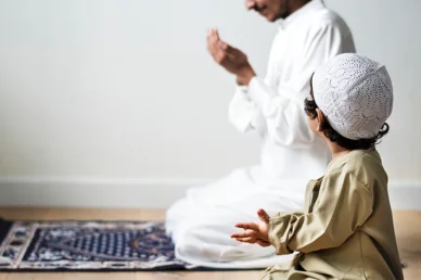 القدوة والتربية الإيمانية: أب وابنه في لحظات الدعاء طفل مسلم يرتدي قبعة صلاة (كوفية) ينظر إلى والده الذي يجلس خلفه على سجادة الصلاة ويرفع يديه بالدعاء في المنزل