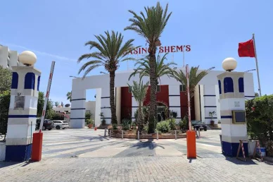 كازينو شمس في أكادير: وجهتك السياحية والترفيهية البارزة المدخل الرئيسي لكازينو شمس في مدينة أكادير نهاراً مع أشجار النخيل والعلم المغربي