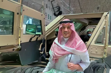 صورة لمسؤول أو ممثل يرتدي الزي العربي التقليدي مبتسماً أثناء استعراض مركبة مدرعة ضخمة ذات لون رملي في معرض أو مؤتمر متخصص بالدفاع والأمن.