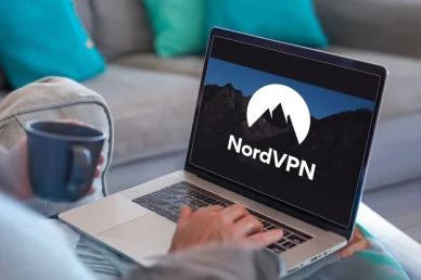 شخص يسترخي على أريكة ممسكاً بكوب، ويستخدم حاسوباً محمولاً تظهر على شاشته علامة NordVPN.