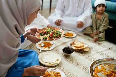 عائلة مسلمة تتناول وجبة طعام معاً امرأة محجبة تقدم السلطة أثناء تناول عائلة مسلمة وجبة تقليدية على الأرض مع التمر والأرز.
