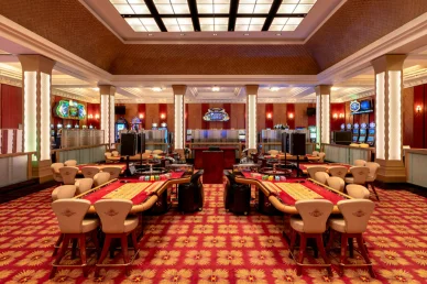 صالة ألعاب كازينو فسيحة وفاخرة تتميز بطاولات الروليت وألعاب الورق الكلاسيكية المغطاة بالقماش الأحمر، ومحاطة بكراسي مريحة. تحتوي الغرفة على أعمدة مضيئة أنيقة، وسجاد أحمر منقوش، وماكينات قمار تظهر في الخلفية.