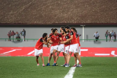 مجموعة من لاعبي المنتخب المصري بقمصانهم الحمراء والبيضاء يعانقون بعضهم البعض بحماس احتفالاً بهدف على أرض الملعب.