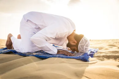 الخشوع في الخلاء: سجدة طمأنينة وسط رمال الصحراء صورة لرجل مسلم يرتدي ثوباً أبيض وقبعة صلاة (كوفية) في حالة سجود على سجادة صلاة زرقاء فوق رمال الصحراء تحت أشعة الشمس الدافئة