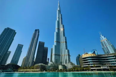 منظر نهاري مهيب لبرج خليفة الشاهق في وسط مدينة دبي، يبرز تصميمه المعماري الزجاجي مع مياه النافورة والمباني المحيطة تحت سماء زرقاء صافية.