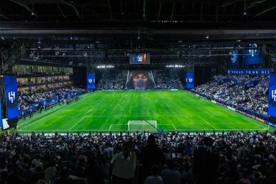 تيفو الهلال المرعب: لوحة فنية وجماهيرية استثنائية في الملعب لقطة واسعة لملعب مغلق وممتلئ بجماهير نادي الهلال، يظهر تيفو عملاق لشخصية مقنعة مرعبة خلف المرمى، وأرضية خضراء مضاءة ولافتات زرقاء تحمل شعار النادي