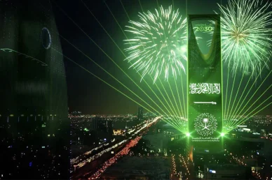برج المملكة في الرياض مضاء باللون الأخضر ليلاً، يعرض علم السعودية وشعار رؤية 2030 مع ألعاب نارية وأضواء ليزر.