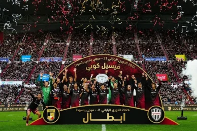 اعبو فريق فيسيل كوبي يحتفلون برفع درع بطولة الدوري الياباني لعام 2024 وسط تساقط القصاصات الملونة في ملعب ممتلئ بالجماهير وتقف أمامهم لافتة كبيرة كتب عليها "أبطال"