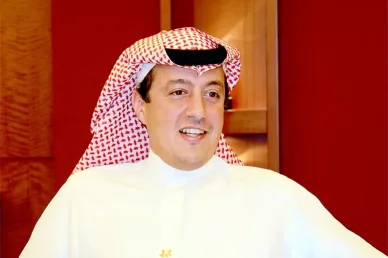 صورة شخصية لمسؤول أو رجل أعمال عربي يبتسم، يرتدي الزي التقليدي (غترة حمراء وبيضاء، عقال أسود، وثوب أبيض) بخلفية خشبية حمراء دافئة.