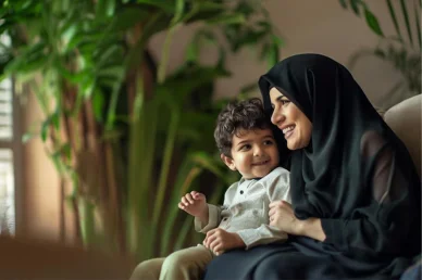 امرأة عربية ترتدي الحجاب تبتسم وتجلس بجانب طفلها الصغير السعيد في أجواء منزلية دافئة مع نباتات خضراء.