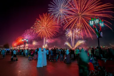 لقطة ليلية لحشد على كورنيش أم القيوين يشاهد عرض الألعاب النارية، بجانب لوحة مضيئة لهيئة السياحة UAQ Tourism.