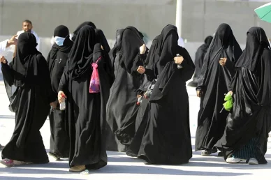 مجموعة من النساء يرتدين العبايات السوداء والنقاب يمشين في الهواء الطلق في يوم مشمس، وإحداهن ترتدي كمامة زرقاء.