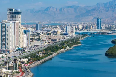 مشهد بانورامي لمدينة رأس الخيمة يضم أبراجاً على الكورنيش، غابات القرم، والمياه الزرقاء مع جبال الحجر في الخلفية.