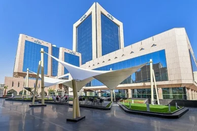 مجمع مباني المقر الرئيسي للبنك العربي الوطني ويحمل علامة (anb) على واجهاته الزجاجية، مع ساحة خارجية واسعة تبرز فيها مظلات بيضاء بتصميم عصري وأماكن للجلوس تحت سماء زرقاء صافية.