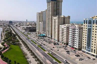 منظر جوي لشارع الكورنيش في مدينة إماراتية يضم أبراجاً سكنية عالية ومواقف سيارات ومساحات خضراء محاذية للمياه.