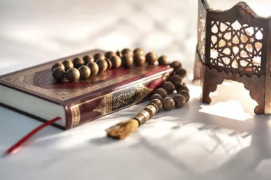 القرآن الكريم والذكر: لحظات من السكينة والروحانية الإسلامية نسخة من القرآن الكريم مع مسبحة خشبية وفانوس تقليدي تضيئها أشعة الشمس الدافئة في مشهد إسلامي روحاني