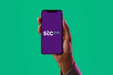 يد تمسك بهاتف ذكي يعرض شعار تطبيق المحفظة الرقمية إس تي سي باي (stc pay) على شاشة أرجوانية بخلفية خضراء.