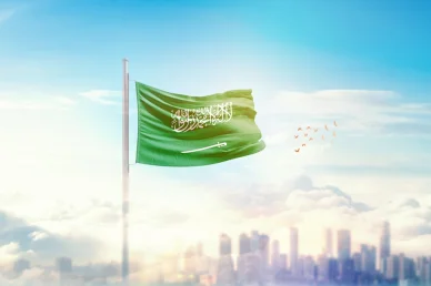 علم المملكة العربية السعودية يرفرف عالياً في سماء زرقاء مليئة بالسحب فوق أفق مدينة حديثة ضبابية مع طيور تحلق.