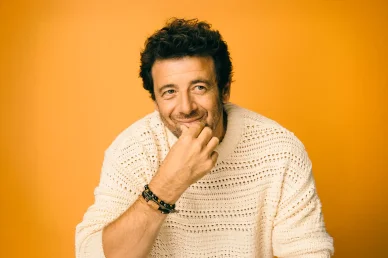 Patrick Bruel souriant, vêtu d’un pull beige sur fond orange.