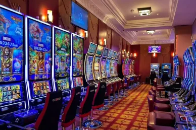 صف طويل من أحدث ماكينات القمار (السلوتس) المضيئة بشاشات ملونة وعالية الدقة داخل صالة كازينو فاخرة، مع سجاد أحمر منقوش أنيق وكراسي مريحة للاعبين.