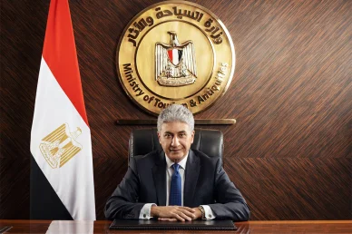وزارة السياحة والآثار في جمهورية مصر العربية مسؤول يجلس على مكتبه ببدلة رسمية، وخلفه جدار خشبي مزين بلوحة ذهبية بارزة تحمل علامة وزارة السياحة والآثار، وإلى جانبه علم جمهورية مصر العربية.