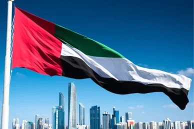 علم دولة الإمارات العربية المتحدة يرفرف عالياً أمام أفق مدينة حديثة تضم ناطحات سحاب تحت سماء زرقاء صافية.