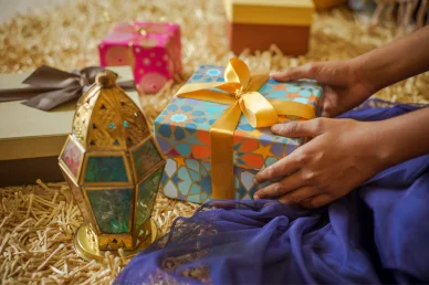 تجهيز هدايا رمضان وعيد الفطر المبارك يدان تمسكان بصندوق هدية مزين بزخارف إسلامية وشريط أصفر، بجوار فانوس رمضان تقليدي وهدايا أخرى على سجادة.