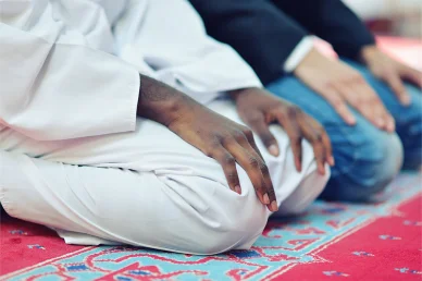 الخشوع والتآخي: لحظات الجلوس للتشهد في الصلاة لقطة مقربة لرجلين مسلمين يجلسان في وضعية التشهد أثناء الصلاة على سجادة حمراء مزخرفة داخل المسجد، أحدهما يرتدي ثوباً أبيض والآخر يرتدي ملابس عصرية