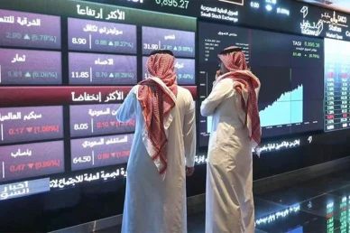 متابعة حركة الأسهم: مستثمرون في السوق المالية السعودية تداول رجلان بالزي السعودي التقليدي ينظران إلى شاشة عرض رقمية كبيرة تظهر بيانات وأسهم السوق المالية السعودية تداول
