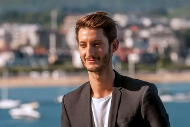 Portrait de l'acteur Pierre Niney souriant légèrement, avec la mer et des bateaux en arrière-plan.