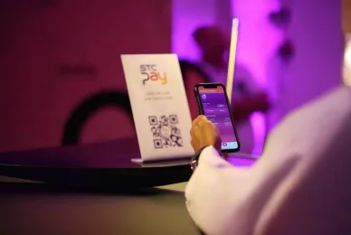 شخص يستخدم هاتفاً ذكياً لإجراء عملية دفع عبر مسح رمز الاستجابة السريعة (QR) المعروض على حامل صغير يحمل علامة (stc pay) وسط إضاءة محيطية بنفسجية.