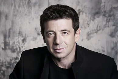 Portrait de Patrick Bruel en veste noire sur fond gris.
