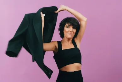 Florence Foresti en tenue décontractée, tenant une veste noire sur un fond rose, avec un air enjoué et dynamique.