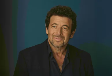 Portrait de Patrick Bruel souriant devant un fond bleu.