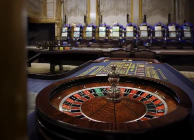 عجلة روليت خشبية كلاسيكية في مقدمة الصورة داخل صالة كازينو أنيقة مع ماكينات قمار في الخلفية.