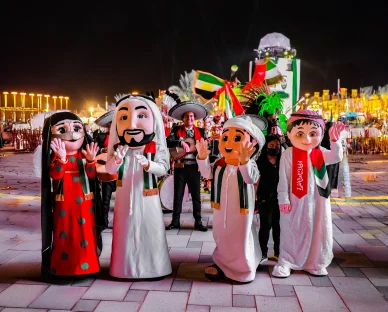 شخصيات كرتونية تمثل العائلة الإماراتية بالزي التقليدي تلوح للجمهور خلال مسيرة احتفالية ليلية.